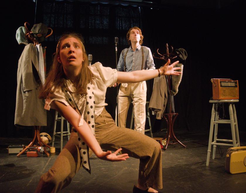 The two actors on stage surrounded by stools, a couple of hatstands and coats, and a 1960s radio. One of the actors is standing with legs and arms outstretched as if holding something very heavy like a boulder.