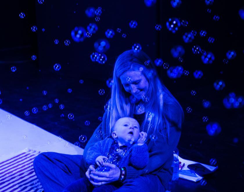 Photo of a parent and their baby enjoying the show, bathed in blue light and surrounded by bubbles.
