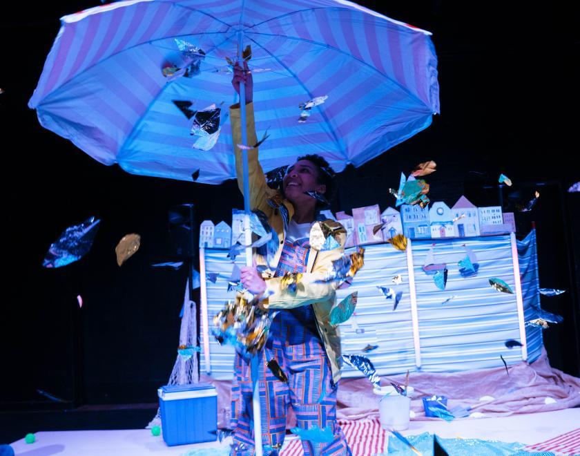 Photo of lead performer standing in the centre of the set holding an open beach umbrella above their head while shiny leaves fall on them.