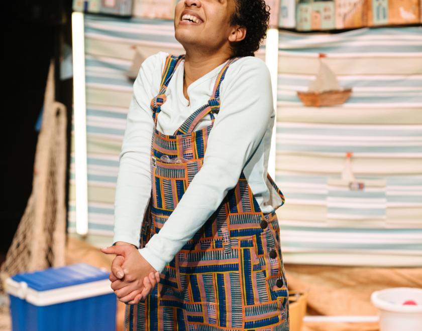 Photo of the lead performer standing in the middle of the set, which is made to resemble a beach, with cool box, towels and backdrop showing a seafront scene with a row of houses and boats visible in the background.