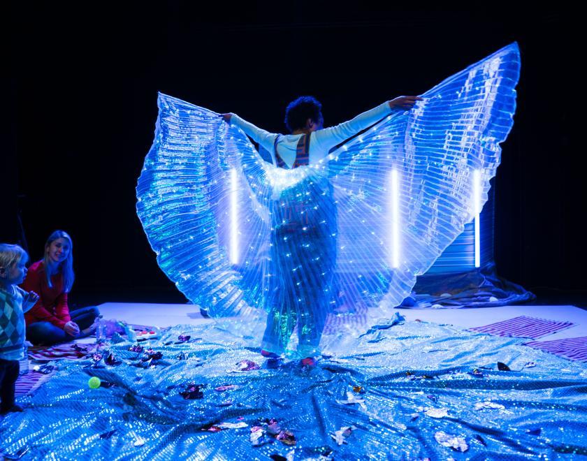 Photo of the lead performer holding a huge piece of light-up see-through fabric behind them like wings, while a baby audience member looks on.