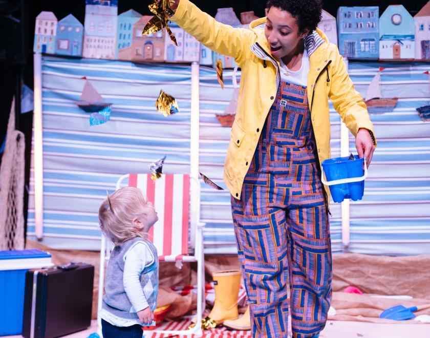 Photo of the lead performer on stage, holding some shiny leaves above a toddler and gently letting them fall over their head