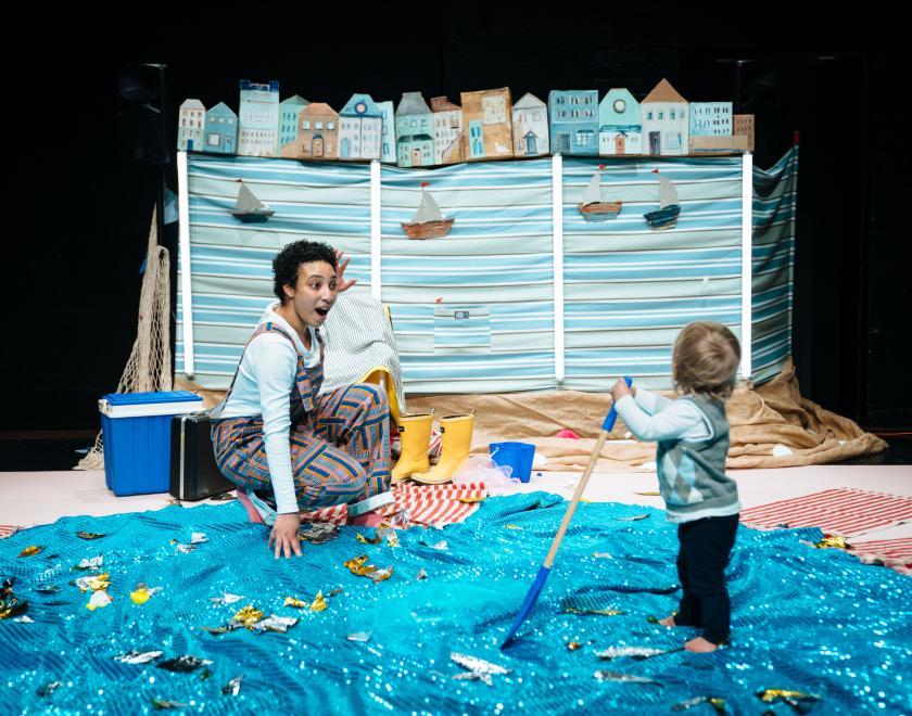 Photo of the lead performer on stage with a toddler, showing them the sea scene backdrop.