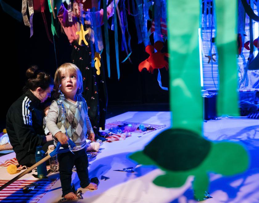 Photo of a toddler audience member standing beneath a large canopy with green and blue streamers and sea creature shapes dangling from it