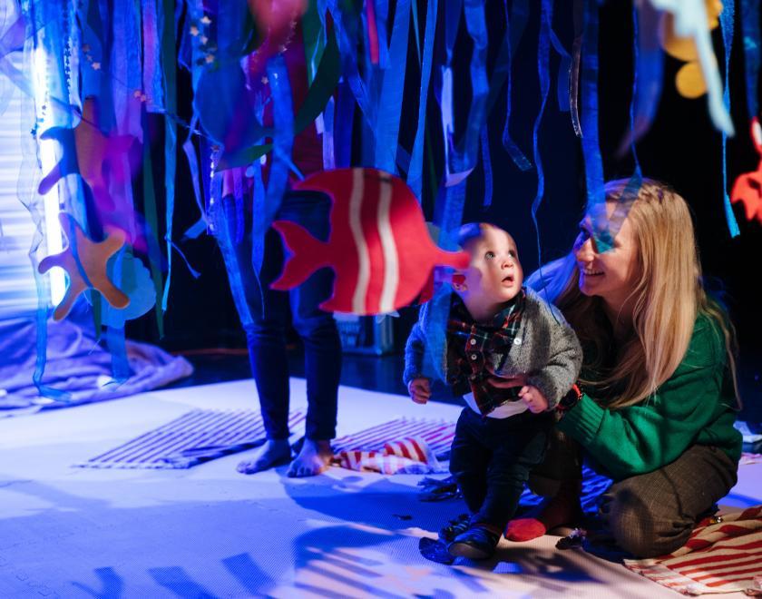Photo of a parent and their toddler beneath the large canopy with blue and green streamers and sea creatures dangling from it.