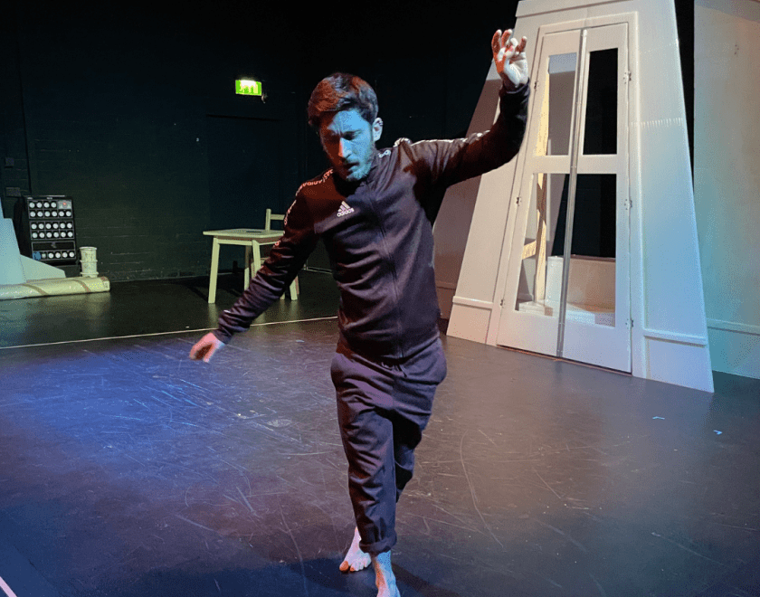 The Dark rehearsal image 4 - a performer stands in the middle of a rehearsal room on tip toes with their arms in the air, as if balancing on a tightrope