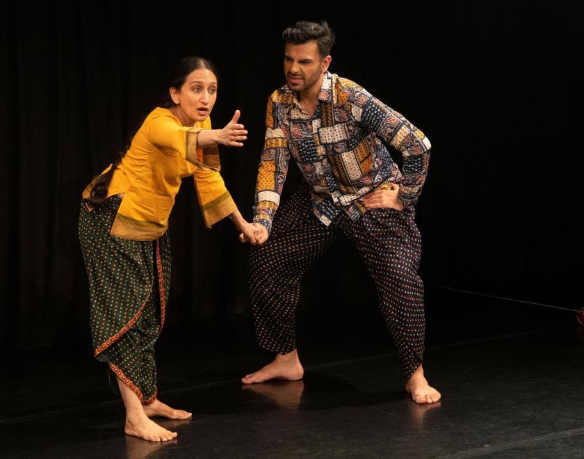 Two performers wearing brightly coloured clothes in front of a black backdrop, holding hands. One is looking out at the audience and gestures a hand towards them, while the other wears a quizzical expression on his face. 