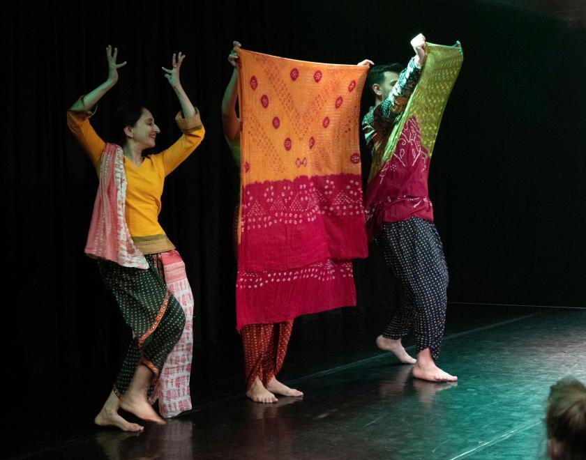 Photo of the performers holding up a bright orange and red scarf and dancing either side of it. 