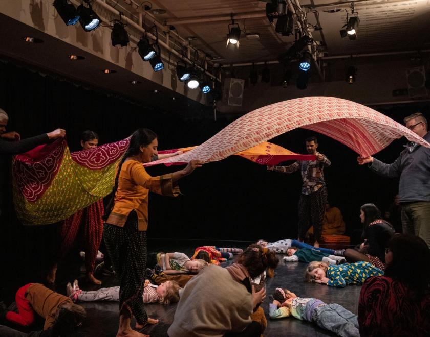 The children in the audience are lying across the floor of the performance space, while the performers and other adults in the audience waft the colourful scarves in the air above them. 