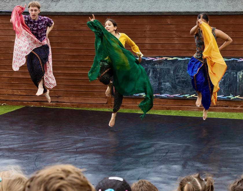 Production photo from an outdoor performance of 'Choogh Choogh', by Vipul Sangoi. Three performers in bright coloured clothes are performing on a large black stage cloth laid on the ground, in front of a barn-like building. The backs of the heads of the audience are visible at the bottom of frame, and the performers are all jumping in the air while holding colourful scarves.