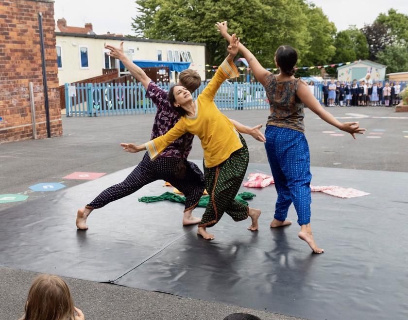 Photo from an outdoor production of Choogh Choogh. Three performers running in a tight circle in the centre of the space, holding their arms out as if imitating an aeroplane.