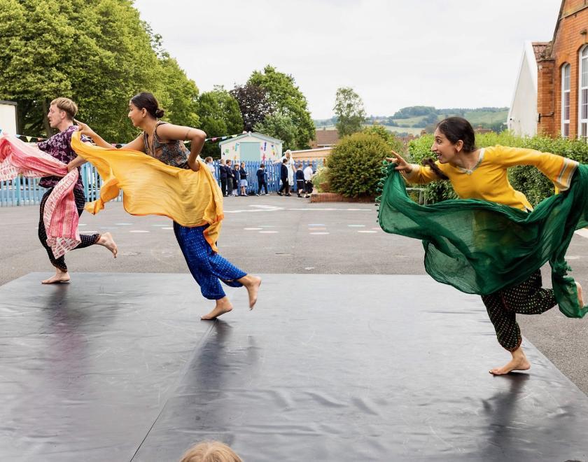 Photo from the same outdoor production. The three performers are in a diagonal line across the space, holding up colourful scarves while they appear to run towards the left of the frame. 