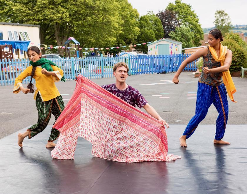Photo from an outdoor production. One of the performers is on their knees holding up a pink scarf, the other two dance on either side of them.