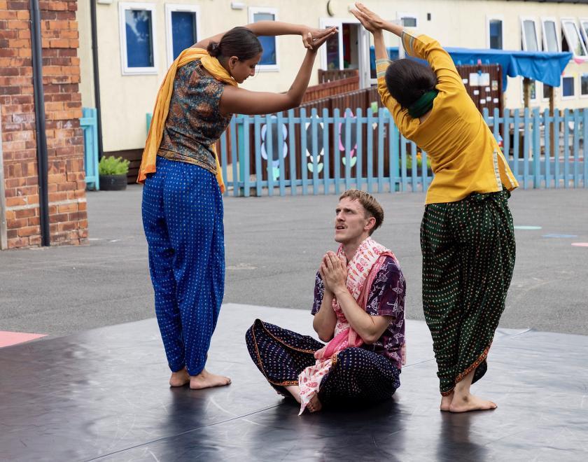 Photo from the outdoor production. One performer sits cross-legged in the centre of the performance space, the other two stand either side of them making an archway above their head with their arms. 