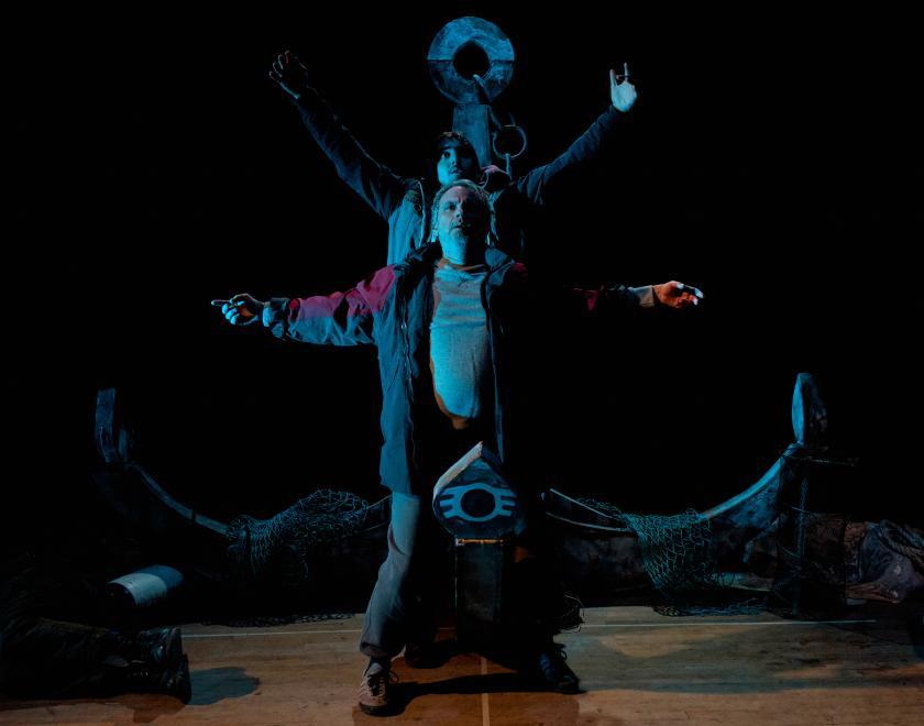 Two performers stand one behind the other, holding their arms up as if in surrender. A large anchor is visible behind them, and the scene is lit by blue stage lighting.