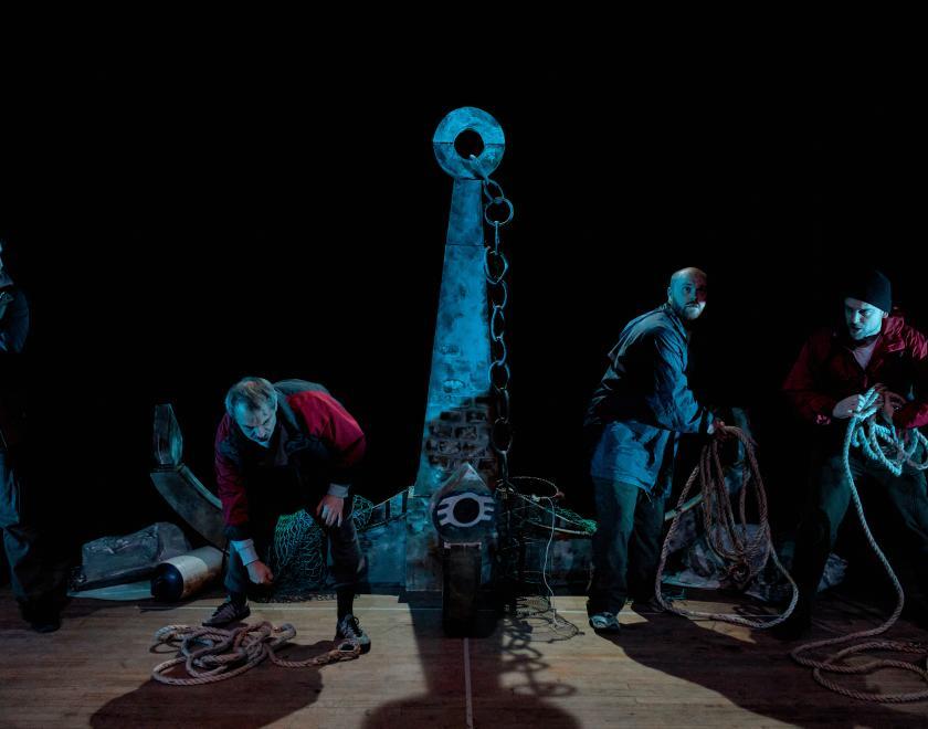 Four performers standing in a row across the stage, each holding a length of rope and moving as if in the act of coiling it up. A large anchor is at the back of the stage, and the whole scene is lit by blue stage lighting. 