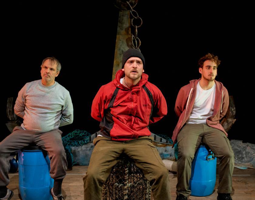 Production photo of 'The Freshwater Five'. Three performers dressed in practical fishing clothing, sit on blue flotation canisters or lobster traps, holding their arms behind their backs as if handcuffed. 