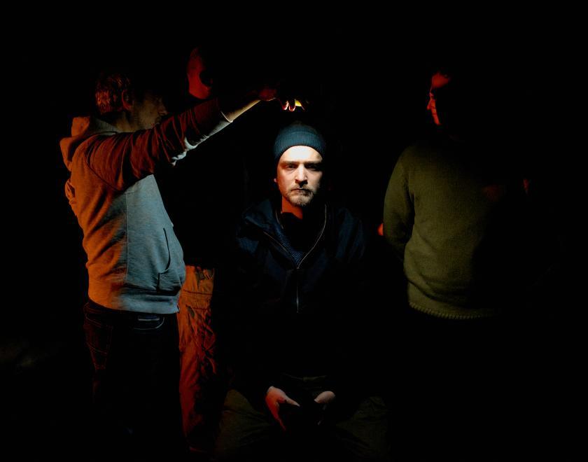 A performer sits on a darkened performance space, illuminated by a small light held above their head by another performer. A third performer is just visible, standing to the right of the seated performer.