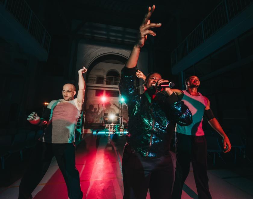 Photo of performer Lady Lykez in a sparkly jacket singing or speaking into a microphone with two performers on either side of her.
