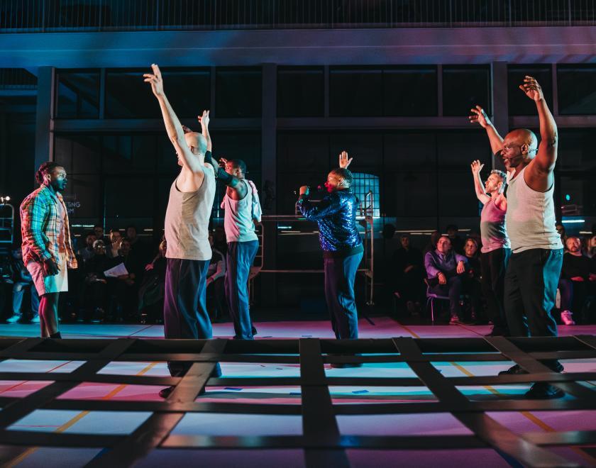Photo of Lady Lykez singing or speaking into a microphone, surrounded by performers with their hands in the air. 