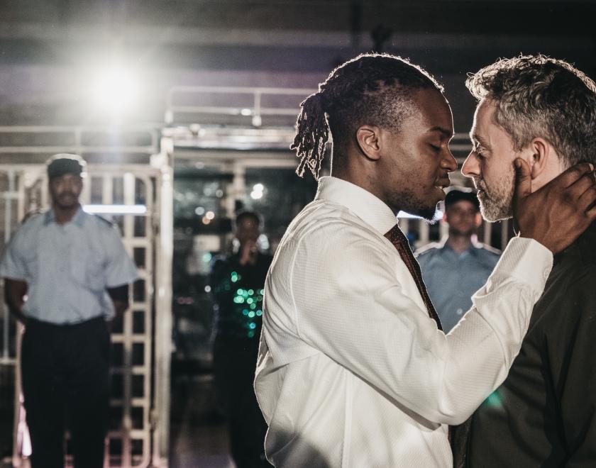 Photo of two performers looking as if they're about to kiss, with a prison guard visible in the background.