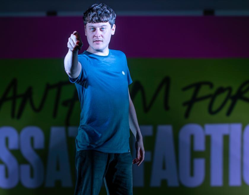 Photo from a performance. An actor on stage pointing towards the audience. A backdrop is visible bearing the text 'Anthem For Dissatisfaction'.