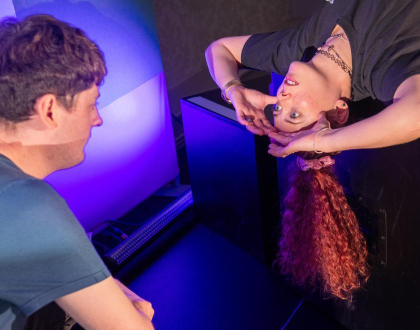 Close-up photo of the two actors, one is lying down over a box with their hands on their head, looking up into the face of the other actor.