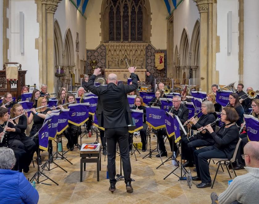 A conductor in black, seen from behind with raised arms, leads Trinity Concert Band during a performance inside a church. Musicians dressed in black play woodwind and brass instruments behind purple Trinity-branded stands, with an audience seated in the foreground and an ornate altar and arches in the background.