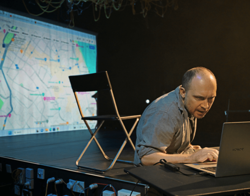 Production photo: A man hunched over a laptop with an empty chair and a projection of a city map 7on the wall behind him.