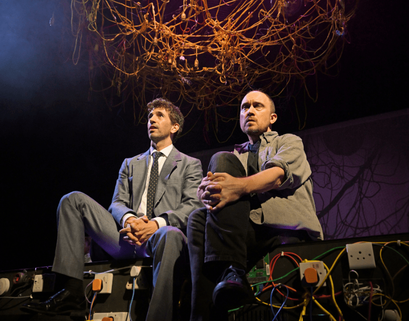 Production photo: Two men sit side by side on the edge of the stage, with a mass of suspended wires visible above them
