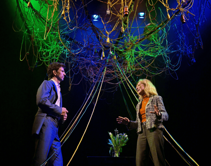 Production photo: A man and a woman stand facing each other with a vase of flowers between them. The wires above them and hanging down are illuminated with blue and green lighting