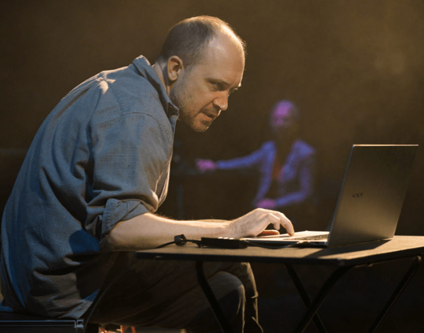 Production photo: a man hunched over a laptop, a woman is visible in the background
