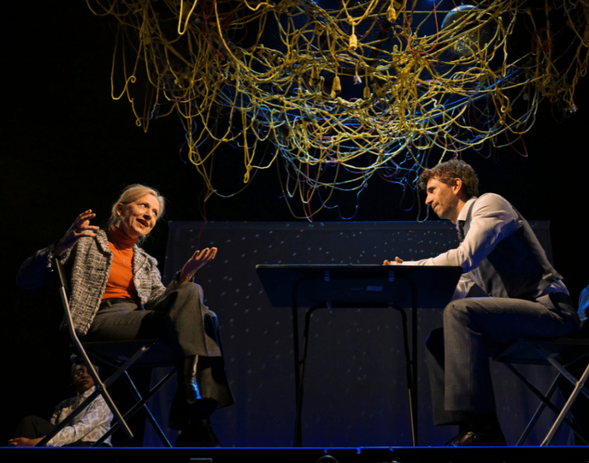 Production photo: A man and a woman having a conversation while sitting at a table. A mass of entangled yellow and blue wires is suspended above them.