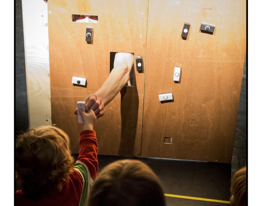 Photo of the wooden box, with the performer's arm stretching out from inside through a hole in the box. Their eyes are visible through another hole. A child in the audience reaches out to press a doorbell held by the performer.
