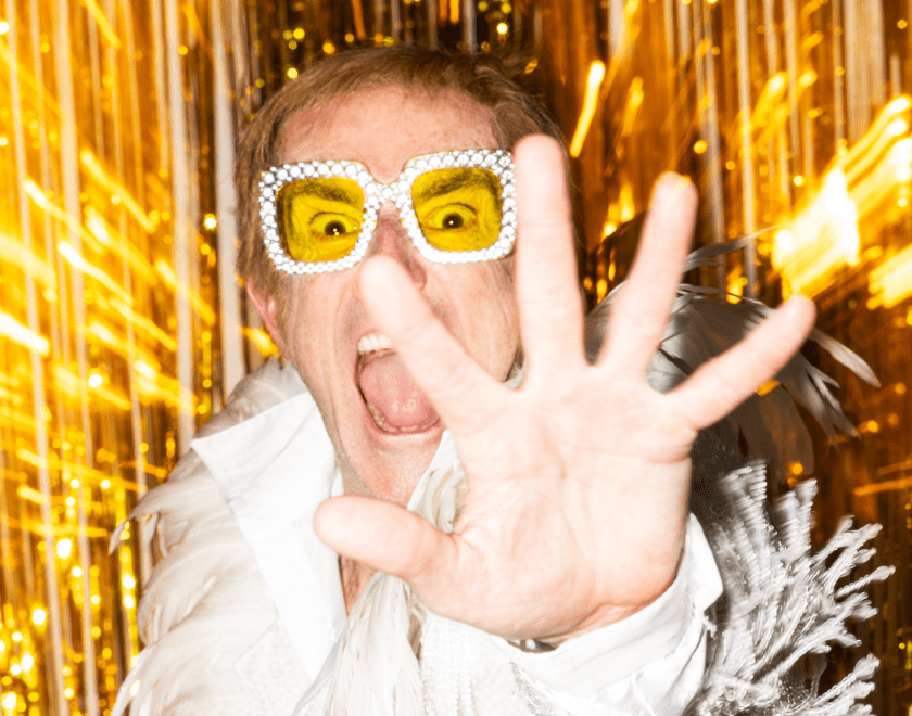 Photo of Henry Naylor dressed as Elton John holding his hand towards the camera as if trying to avoid the paparazzi, against a gold backdrop.