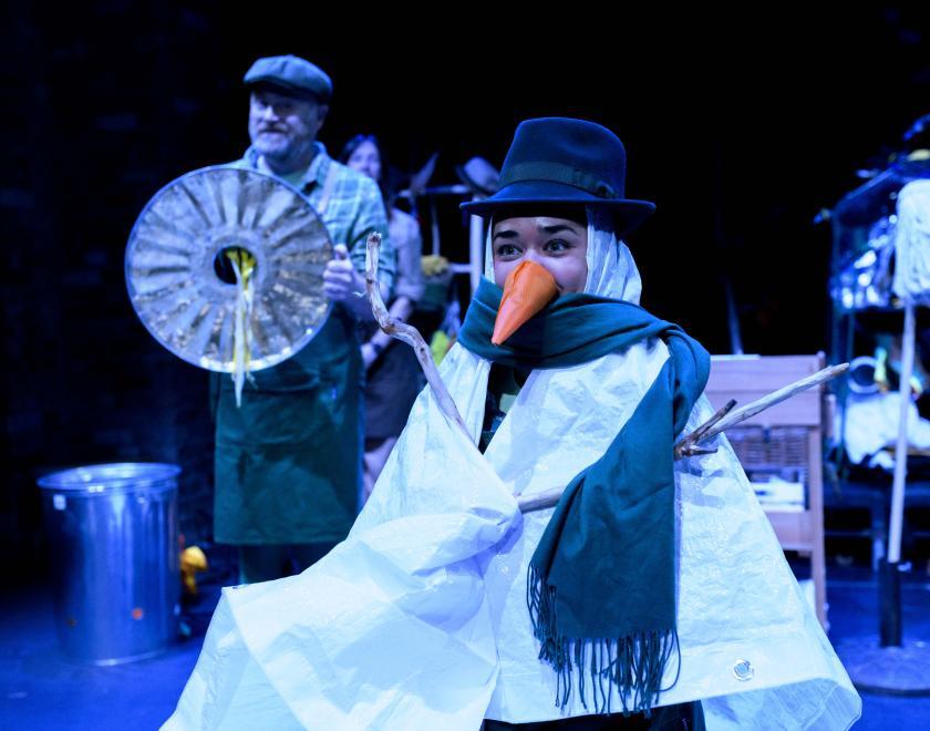 Photo of a performer dressed as a snowman with a hat, scarf and carrot nose, holding two sticks for arms.
