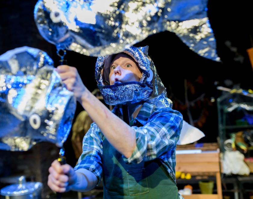 photo of a performer wearing a checked shirt and green dungarees, with a hat in the shape of a silver fish on their head. They're holding two large puppets in the shape of silver fish. 