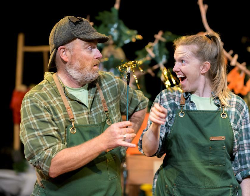 Photo of two performers wearing checked shirts and green dungarees, each holding a small stick with gold streamers coming out the end. 