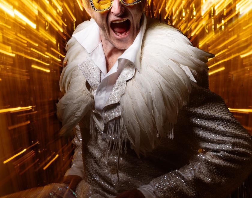 Photo of Henry Naylor dressed as Elton John in front of a gold backdrop, posing as if he's yelling at the camera