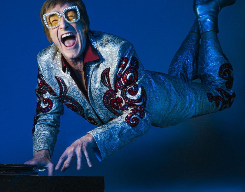 Photo of Henry Naylor dressed as Elton John, playing a piano in front of a blue backdrop. His legs are elevated behind him in the 'Rocketman' pose.