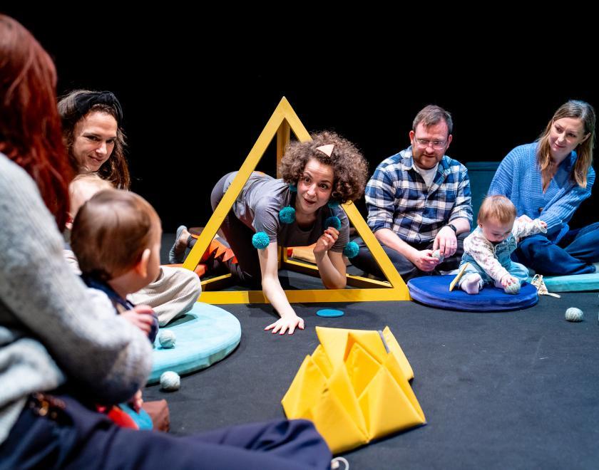 Photo of the performer crawling through a yellow pyramid-shaped frame, surrounded by a audience of adults and babies.