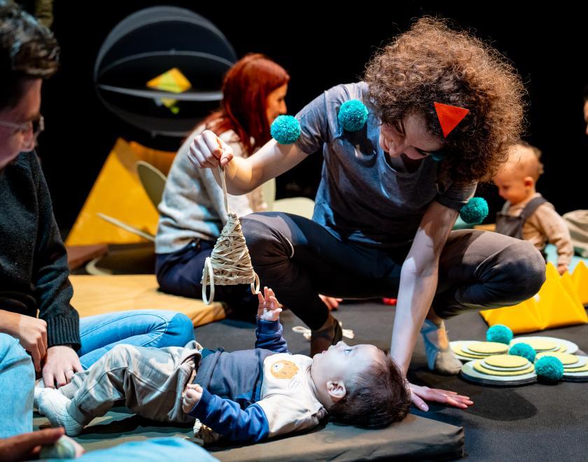 Photo of the performer interacting with a baby, who is lying on one of the soft pads spread around the space. The performer is holding a triangle shaped prop above the baby for them to play with. 