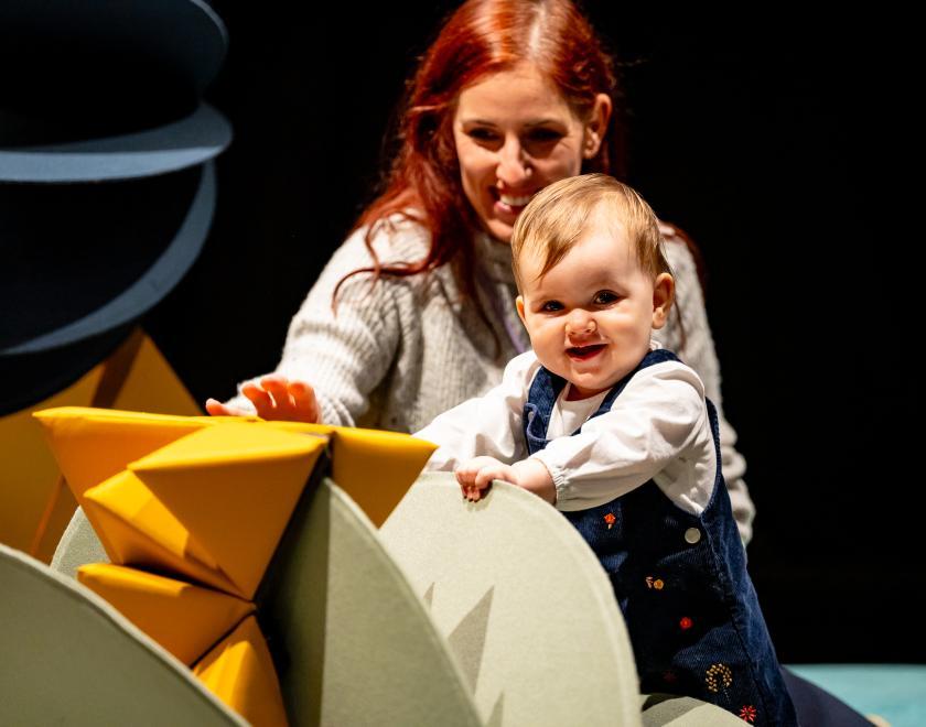 Close-up photo of an adult and baby, both smiling as they interact with a yellow prop shape.