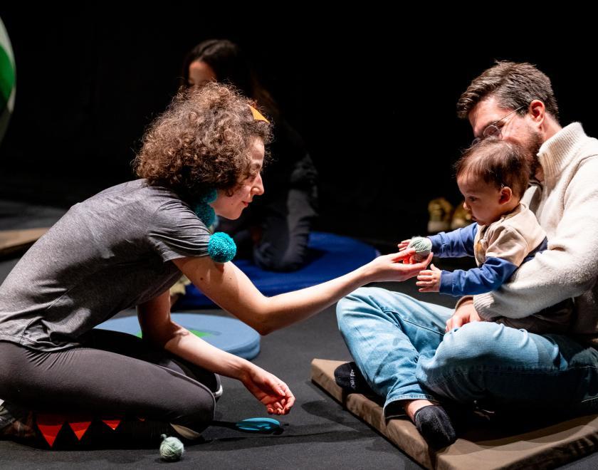 Photo of the performer holding out a ball for baby to play with, while they're being held by their grown-up. 
