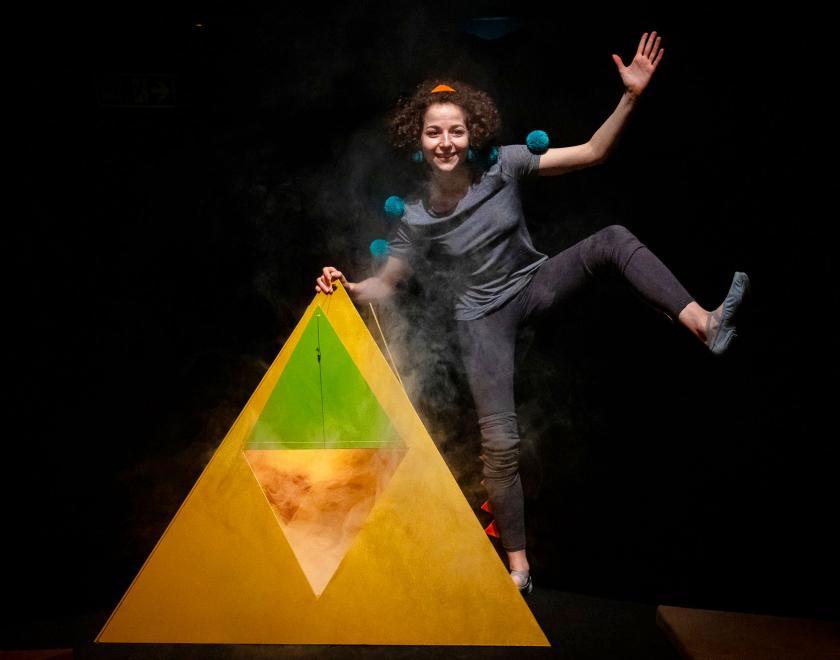 Photo of a performer wearing dark clothes with blue pom-poms sewn onto the sleeves. They are standing next to a large yellow pyramid with an open flap on its front, from which steam or smoke is floating out and up into the air. The performer has one hand on the top of the pyramid, and lifts their other hand and left leg up into the air in a jaunty pose.
