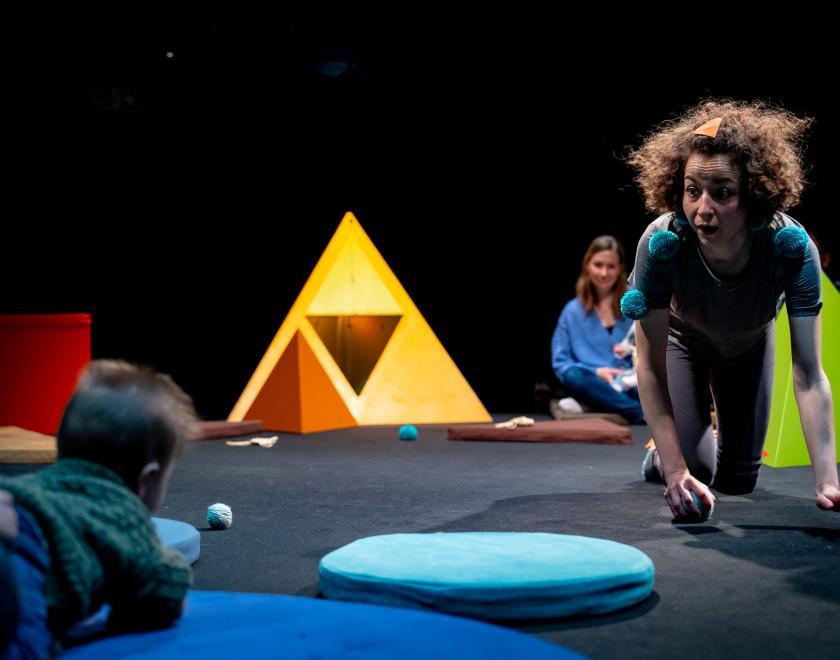 Photo of the performer on hands and knees in the space, as a baby in the audience watches them. The space has various brightly coloured shape props placed around it.