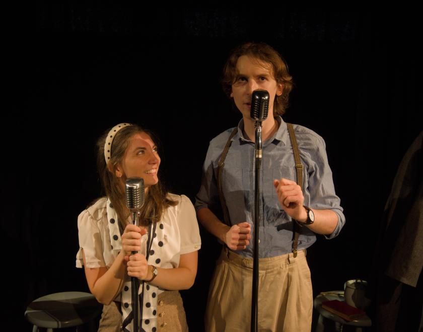 Close-up photo of the actors standing side by side speaking into old fashioned microphones.