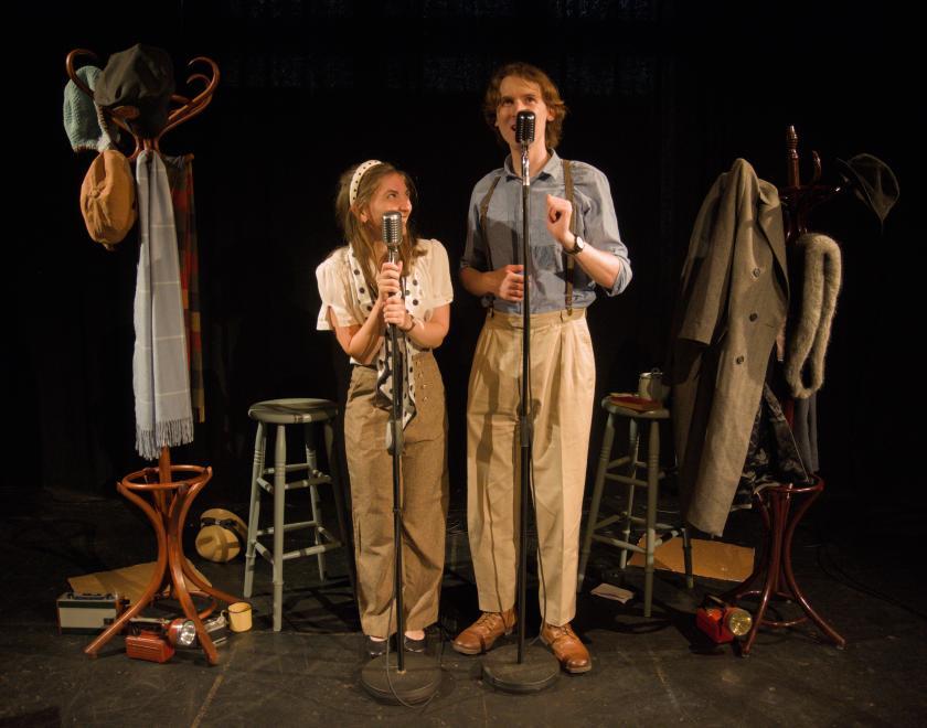 Photo of the two actors standing side by side speaking into microphones, with the rest of the set in the frame.