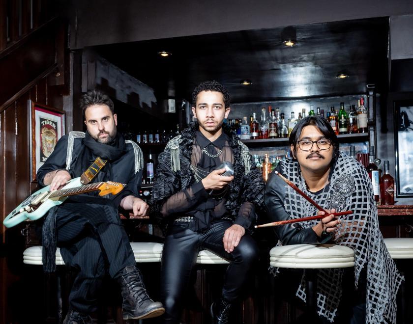 Photo of all three performers sitting or leaning on white bar stools in front of a black bar with a wall of alcohol bottles behind them