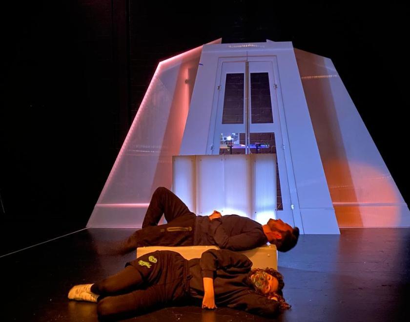The Dark rehearsal image 5 - two performers lie on the floor of a performance space as if asleep, there is a large white structure behind them with a door in the centre